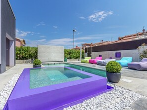 Un área de piscina moderna al aire libre con una piscina rectangular, sillas de descanso coloridas y un muro de hormigón.