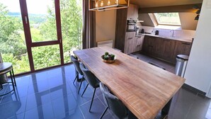 Moderni keittiö, jossa on suuri ruokapöytä, ikkunasta avautuva näkymä vehreään maisemaan ja katosta roikkuva valaisin.