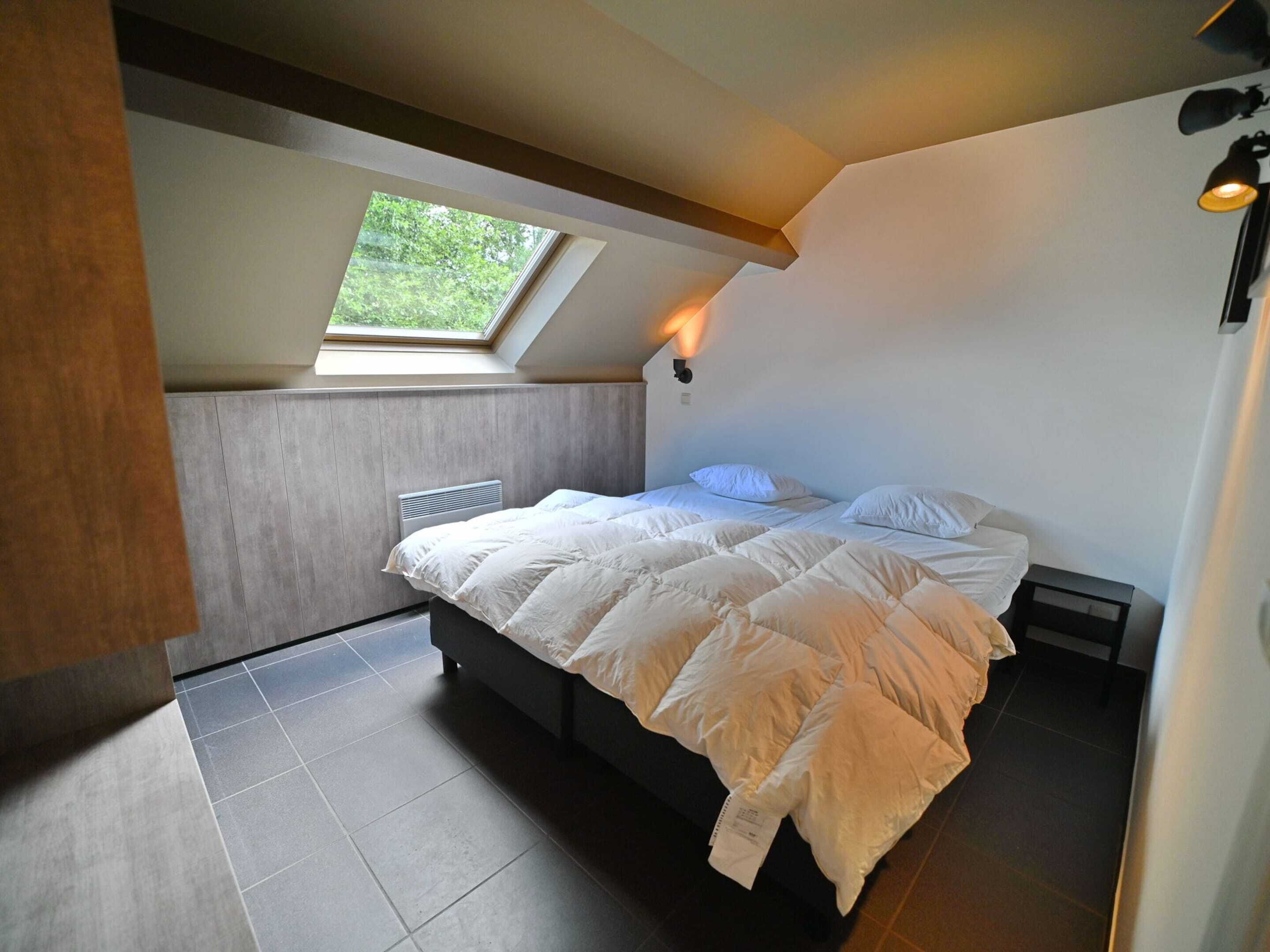 Une chambre avec un puits de lumière, un lit avec une literie blanche, un radiateur et une table de chevet.