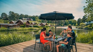 Un groupe de personnes est réuni autour d’une table en bois, sur une terrasse avec vue sur un lac, et des chalets se dressent alentour.