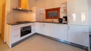 Une cuisine moderne avec des meubles blancs, une cuisinière à gaz, un four, une hotte intégrée et une fenêtre avec des stores.