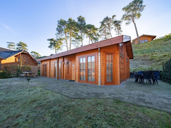 A wooden cabin with large windows, a patio with a table and chairs, and a surrounding grassy area.