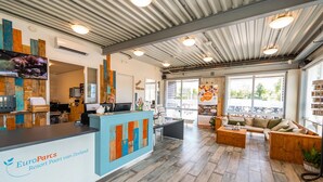A reception area with a wooden reception desk, a seating area with a sofa and coffee table, and a large window providing a view of the outdoors. - Comfortable Chalet With Combi Microwave, Near Rockanje (Hellevoetsluis)