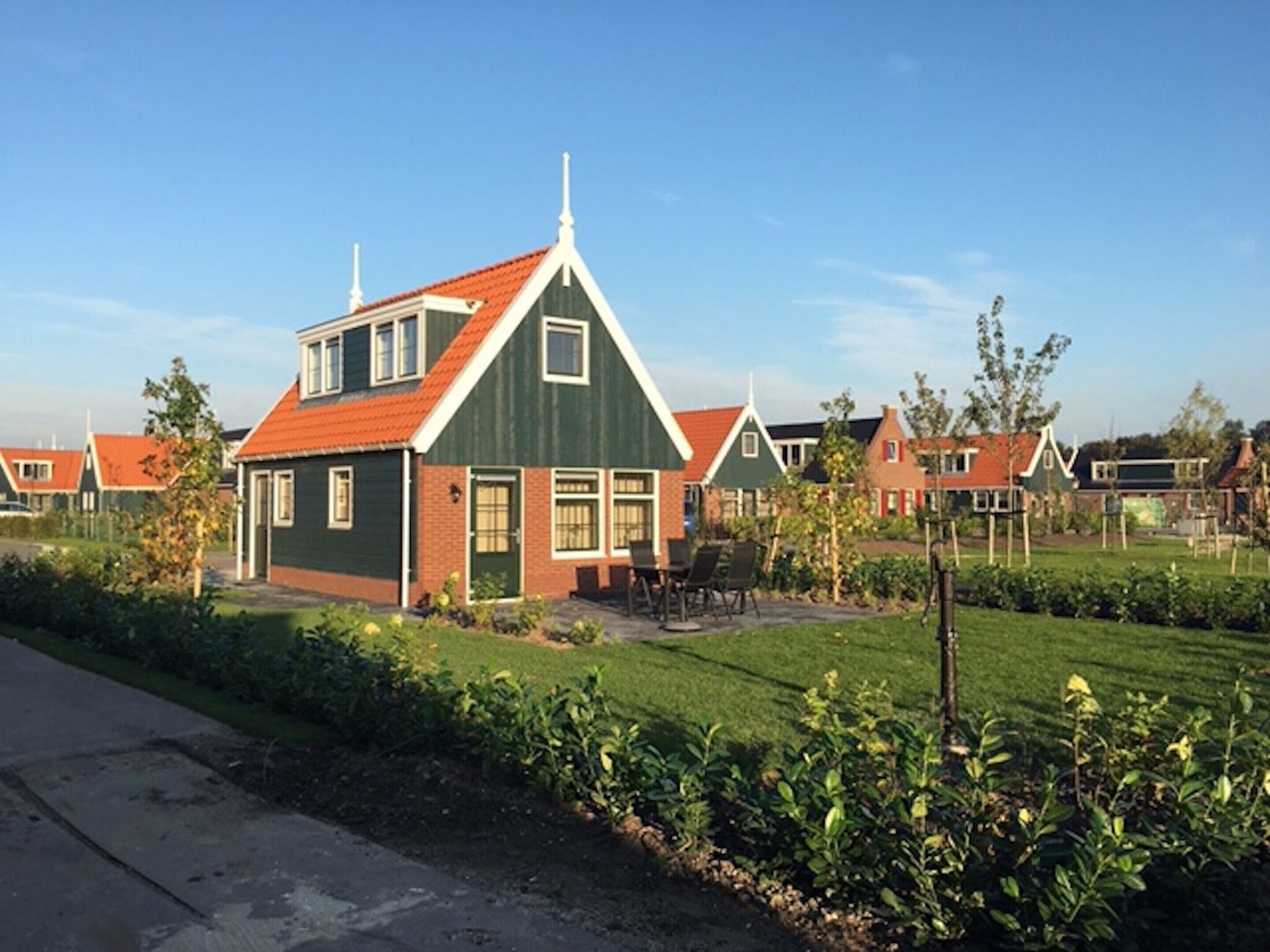 A row of traditional houses with green facades and red roofs, arranged in a residential area with well-maintained lawns and a clear sky.