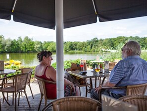 A couple seated at an outdoor table with a view of a lake, surrounded by wicker chairs and a table umbrella. - Chalet With Dishwasher in the Green Kempen (Mol)