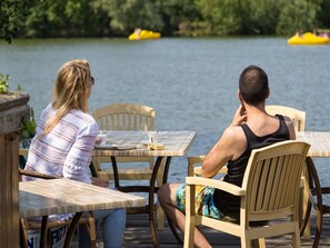 Two people seated at a wooden table with a view of a lake and yellow boats in the background. - Chalet With Dishwasher in the Green Kempen (Mol)