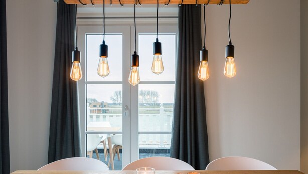 A dining area with a wooden table, four hanging light bulbs, and a view of a body of water through a window. - Modern Apartment 3 Bath Rooms, Close to Lake Veere (Kamperland)