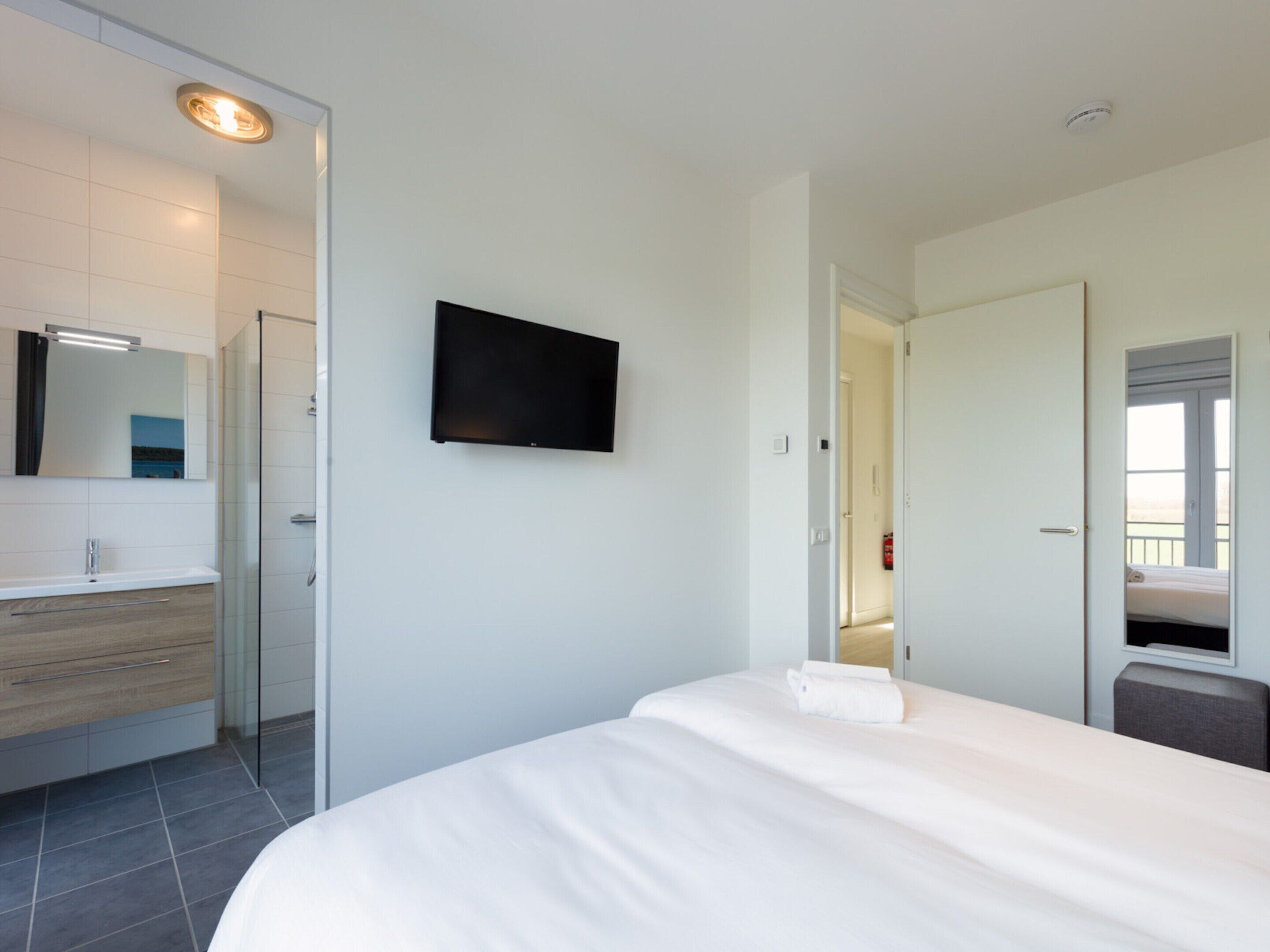 Une chambre d’hôtel moderne dotée d’un grand lit, d’un téléviseur à écran plat fixé au mur, d’une salle de bain séparée avec une douche à parois de verre, et d’un couloir visible qui mène à une autre chambre.
