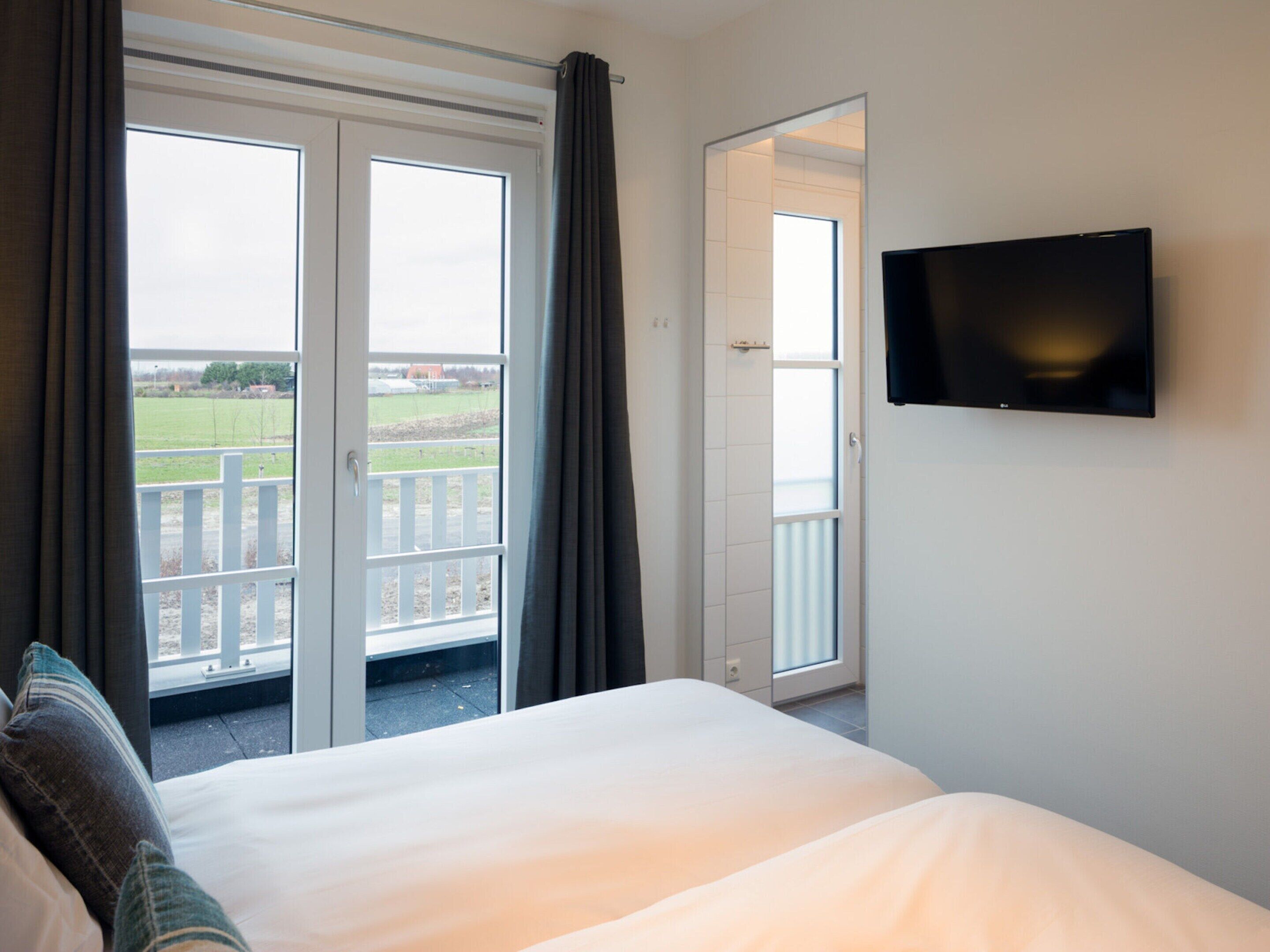 A hotel room with a bed, a television mounted on the wall, and a sliding glass door leading to an outdoor area with a view of a field.