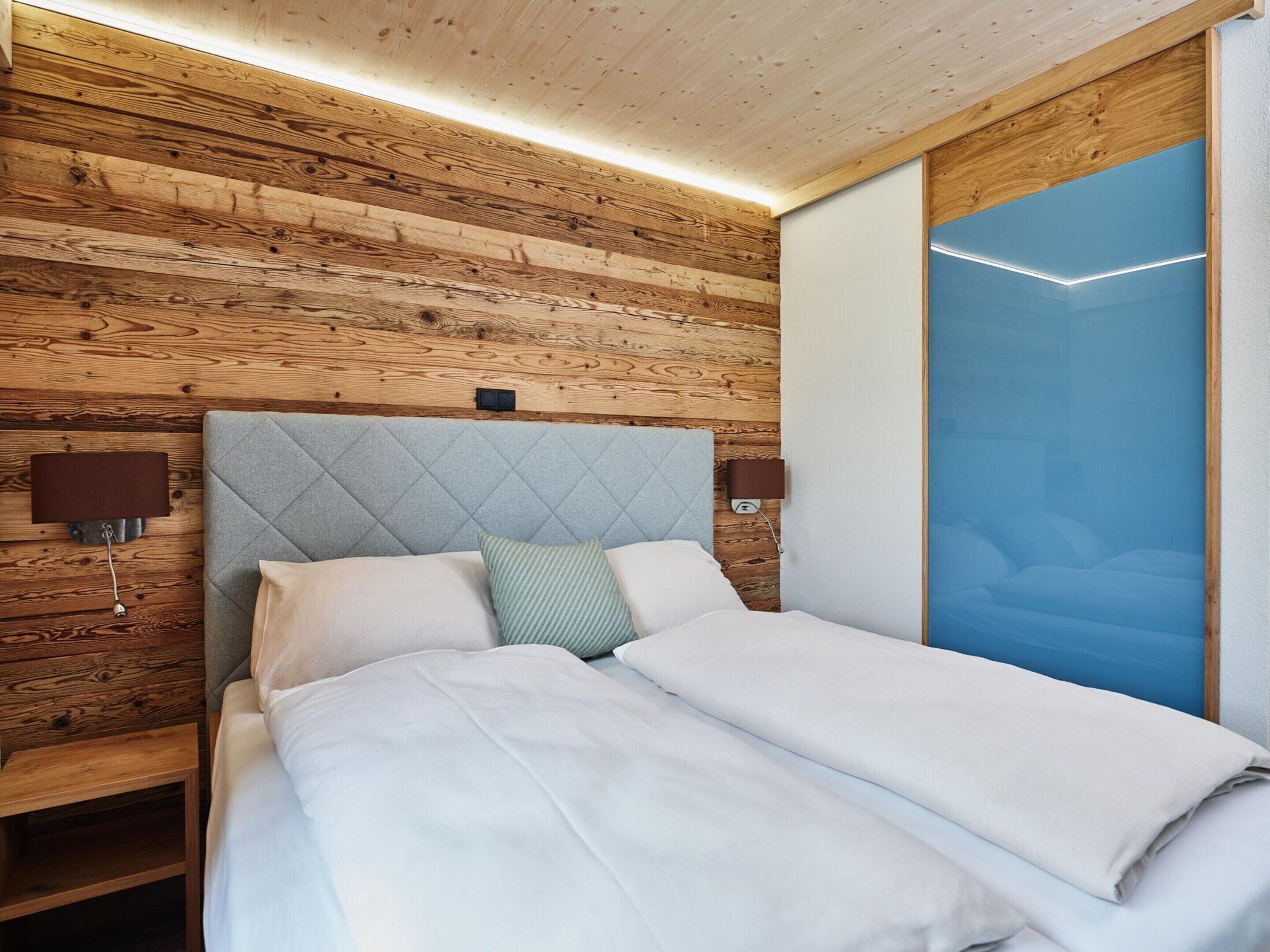 Un dormitorio con panel de pared de madera, cabecero acolchado gris, dos mesitas de noche con lámparas y un ventanal con vista azul.