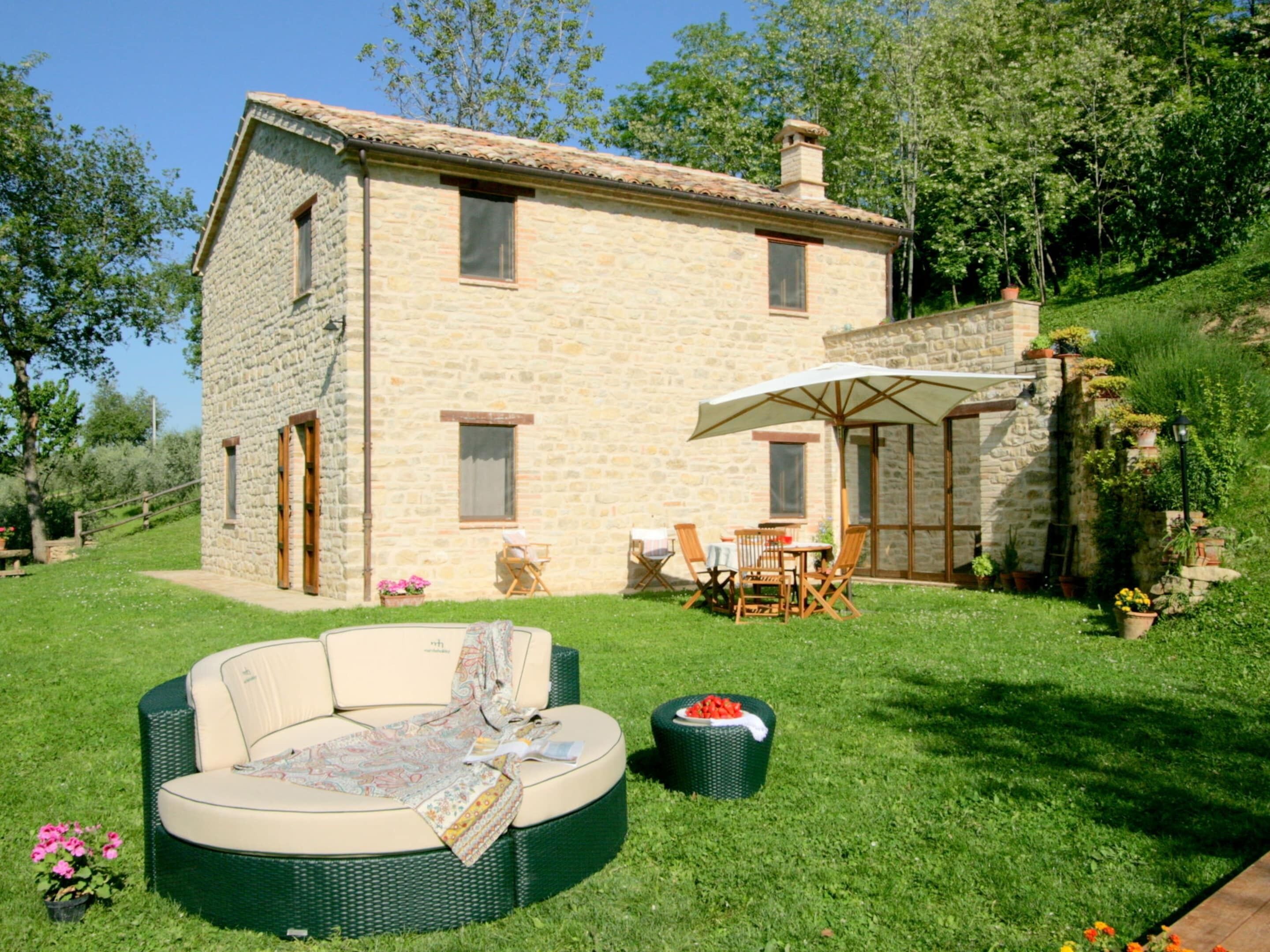 A stone house with a patio area featuring a round wicker sofa, a dining table with chairs, and an umbrella.