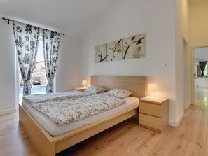 Ein Schlafzimmer mit einem hölzernen Bett, zwei Nachttischen mit Lampen, einem großen floralen Gemälde an der Wand und Blick auf einen Pool durch transparente Vorhänge.