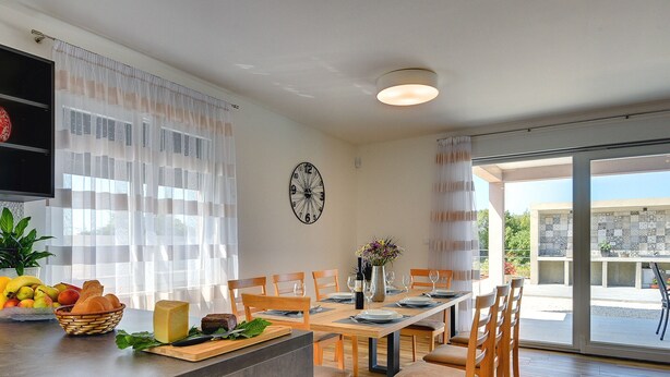 Ein moderner Essbereich mit einem langen Tisch, Stühlen und einer Küchenzeile mit Früchten und Brot.