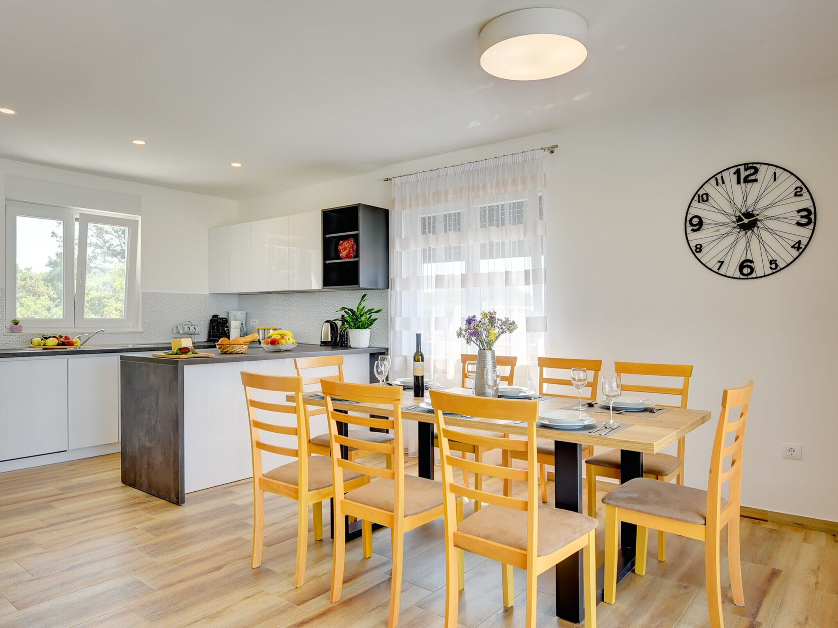 A modern kitchen with a dining area, wooden chairs, a clock on the wall, and a window.