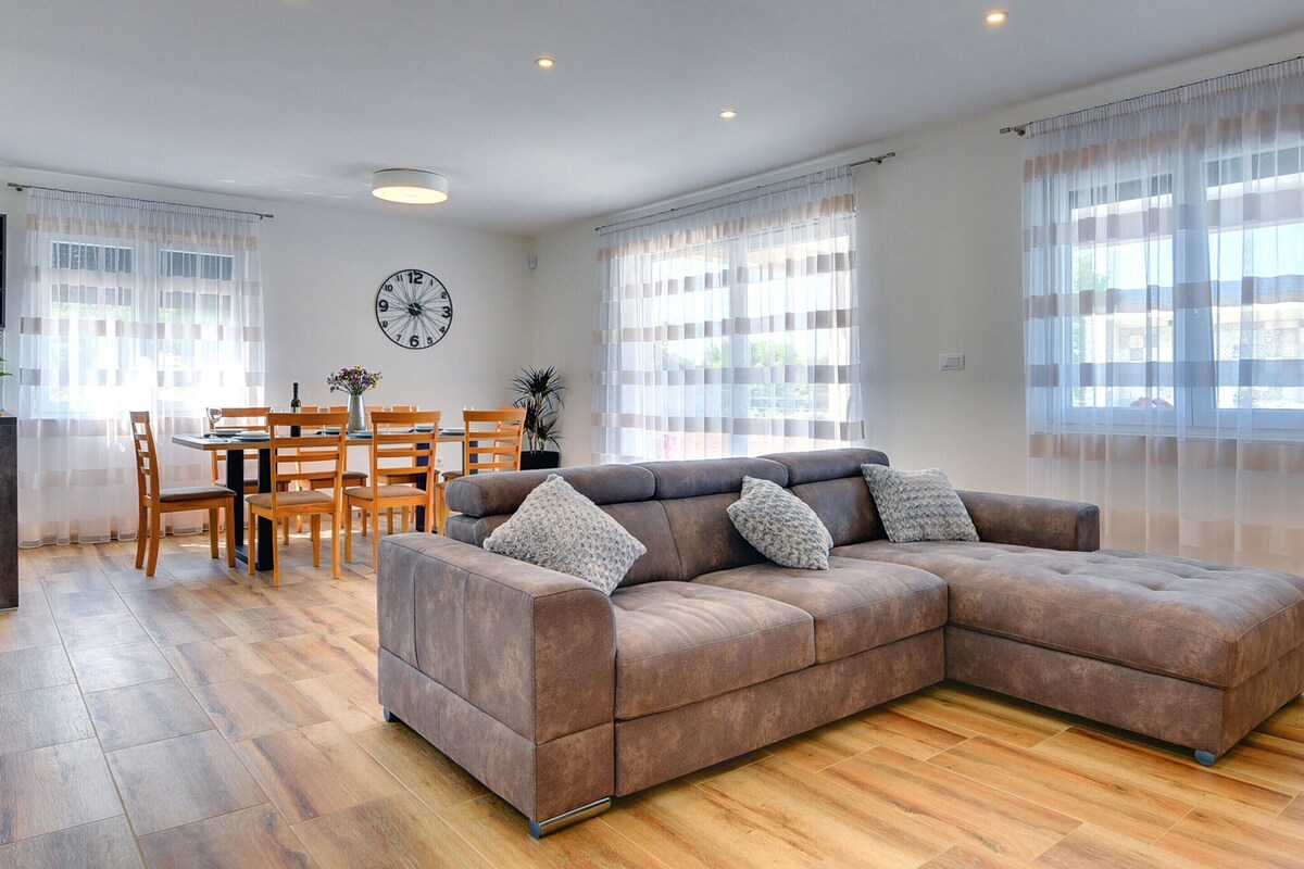 Una sala de estar con un sofá modular, una mesa de comedor de madera, sillas y un reloj en la pared.