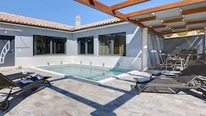 Zona junto a la piscina con sillones de descanso, una parrilla y una pérgola.