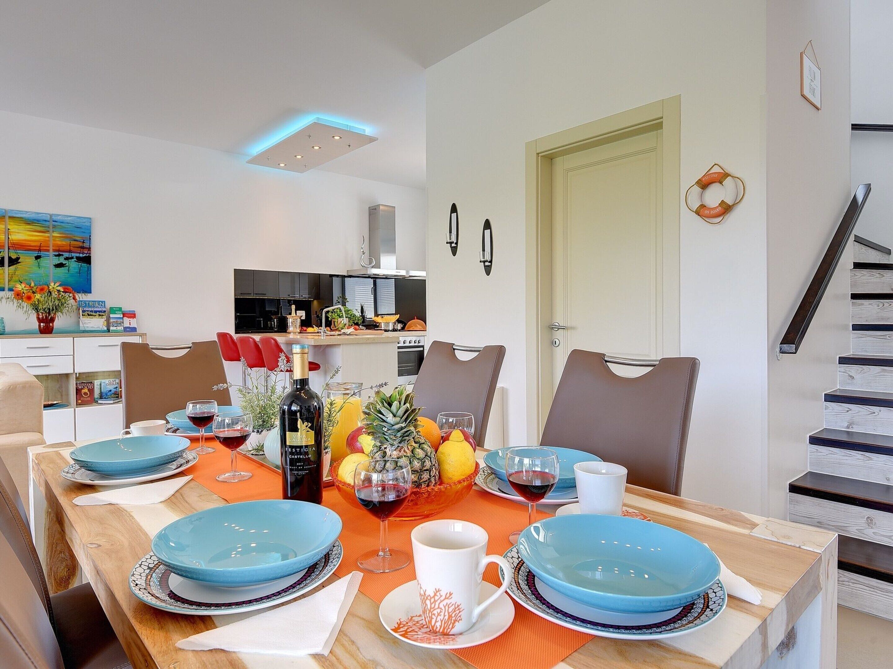 A dining area with a wooden table set for four, blue plates, and a bottle of wine. A kitchen is visible in the background with modern appliances.