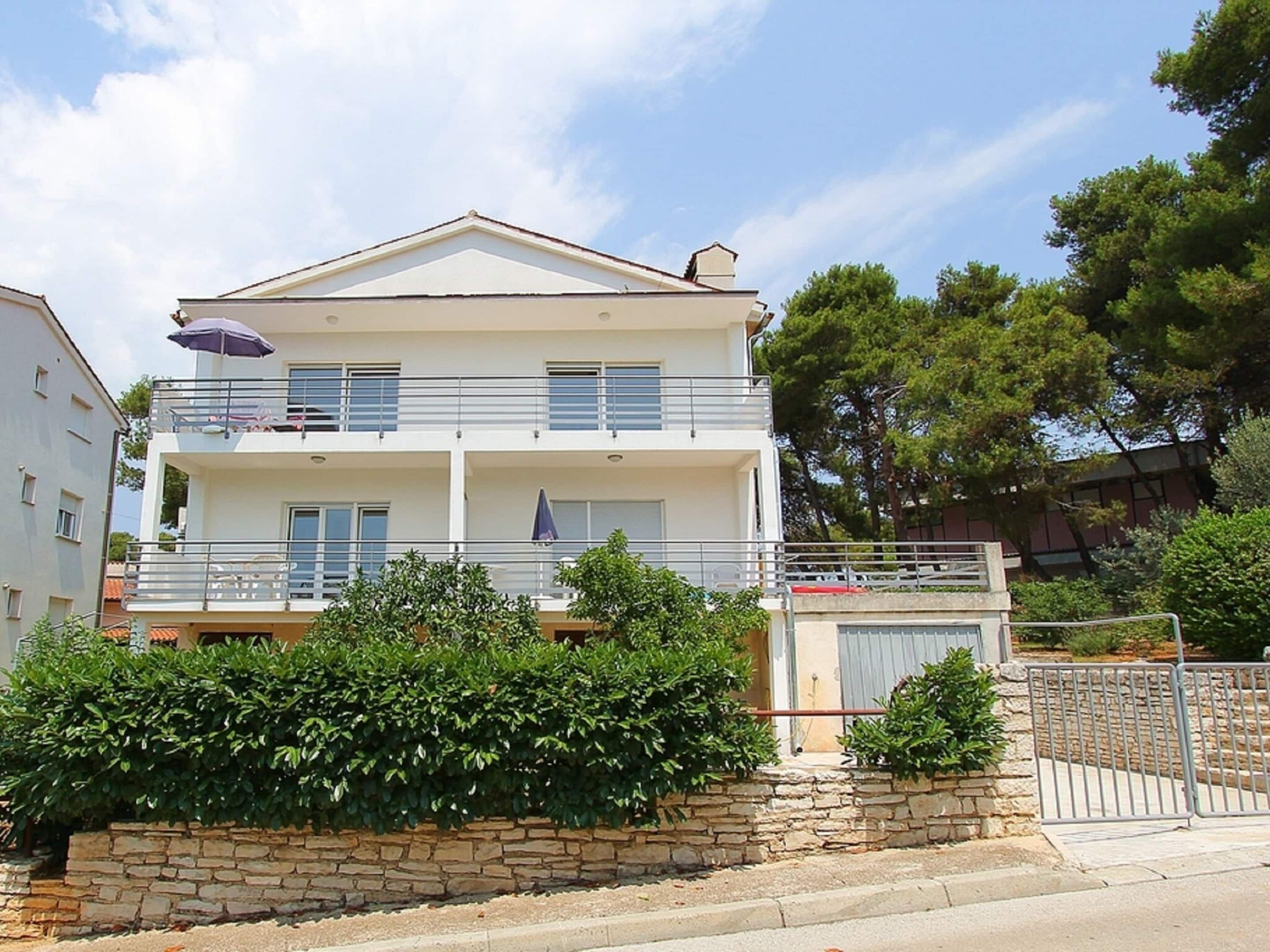 A two-story building with a balcony on the upper floor, a stone wall in front, and a metal gate to the right.