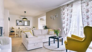 Un salon avec un canapé, un fauteuil jaune, une table basse et, au fond, un coin repas.