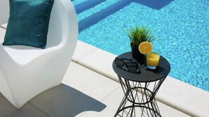 A poolside area with a white chair, a patterned pillow, a small table with a plant, a glass of orange juice, and a pair of sunglasses. - Luxury Apartment With Private Pool, Near the Beach and Close to Pula (Fazana)