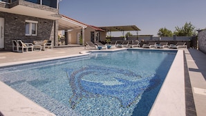 Ein Swimmingpool mit Mosaikfliesen, eine überdachte Terrasse und Liegestühle.