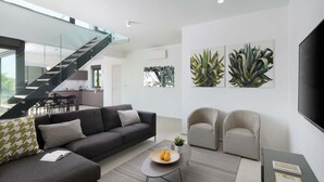 Una sala de estar moderna con un sofá gris, una mesa de centro y dos sillones. Se ve una escalera que conduce a un nivel superior, y hay obras de arte en las paredes.