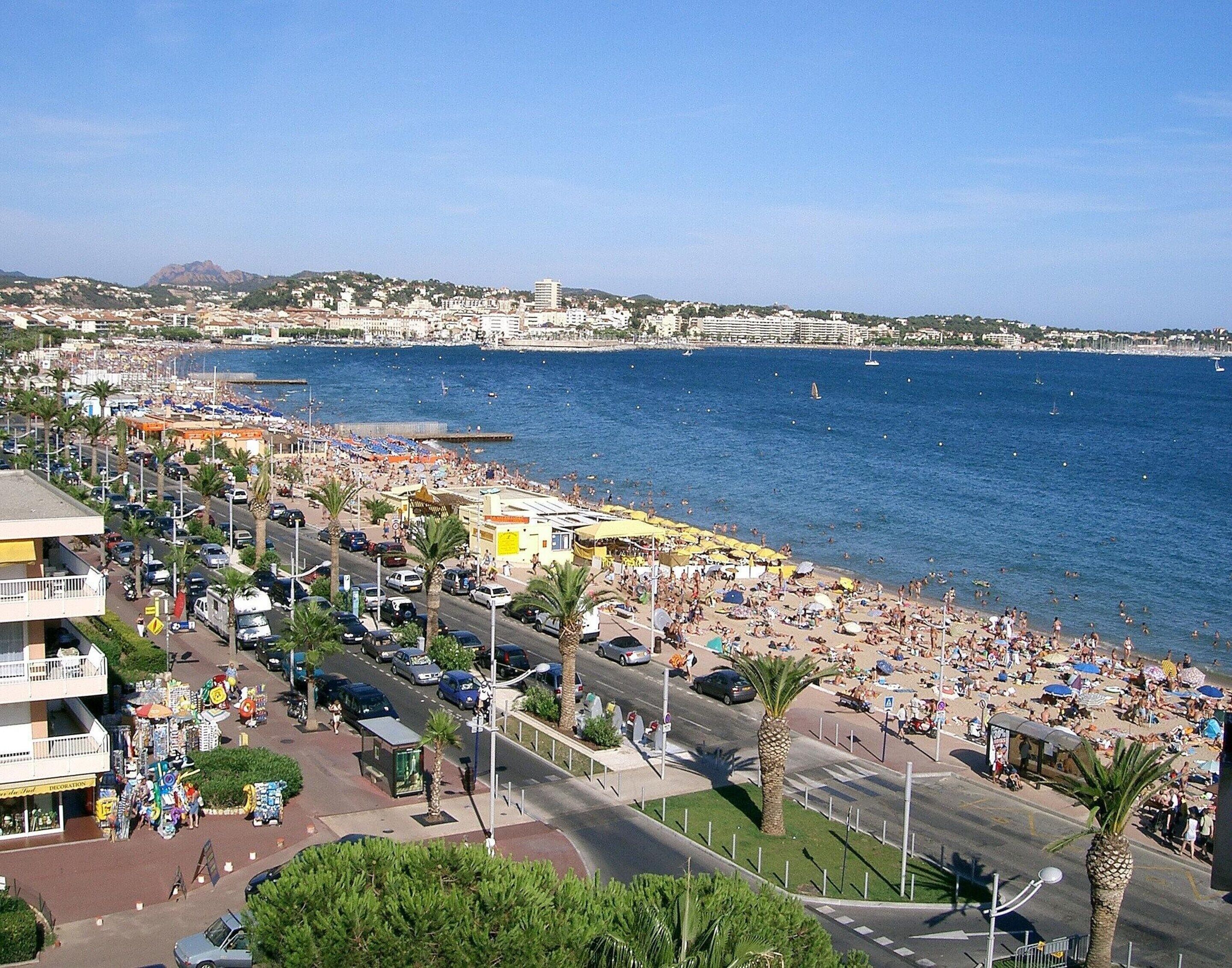 A coastal cityscape with a busy beach, clear blue sea, and a mountainous backdrop.