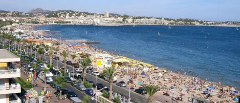 A coastal cityscape with a busy beach, clear blue sea, and a mountainous backdrop.