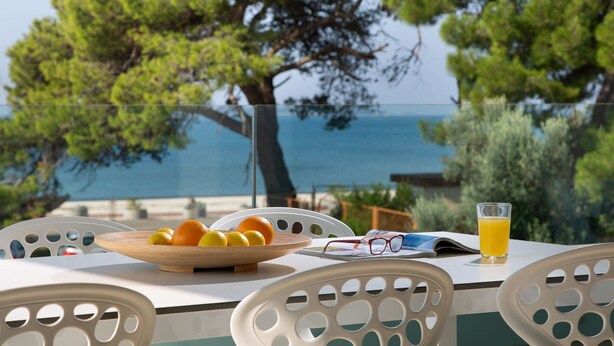 Una mesa con un bol de fruta, un vaso de zumo de naranja y un par de gafas en una terraza con vistas al mar.