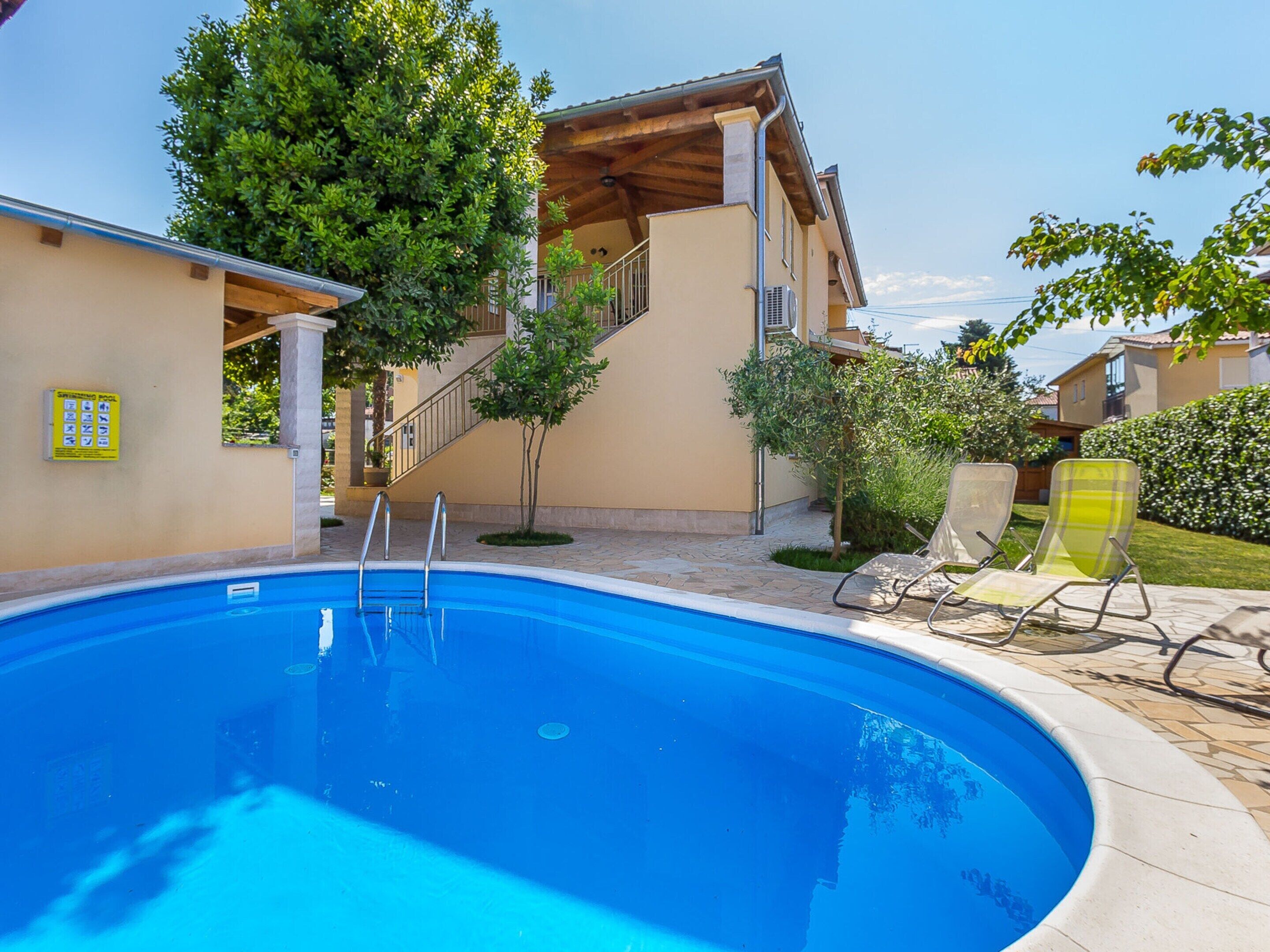 A swimming pool with steps leading to a covered area, surrounded by greenery and a tiled patio.