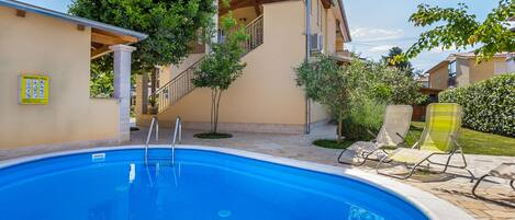 A swimming pool with steps leading to a covered area, surrounded by greenery and a tiled patio.