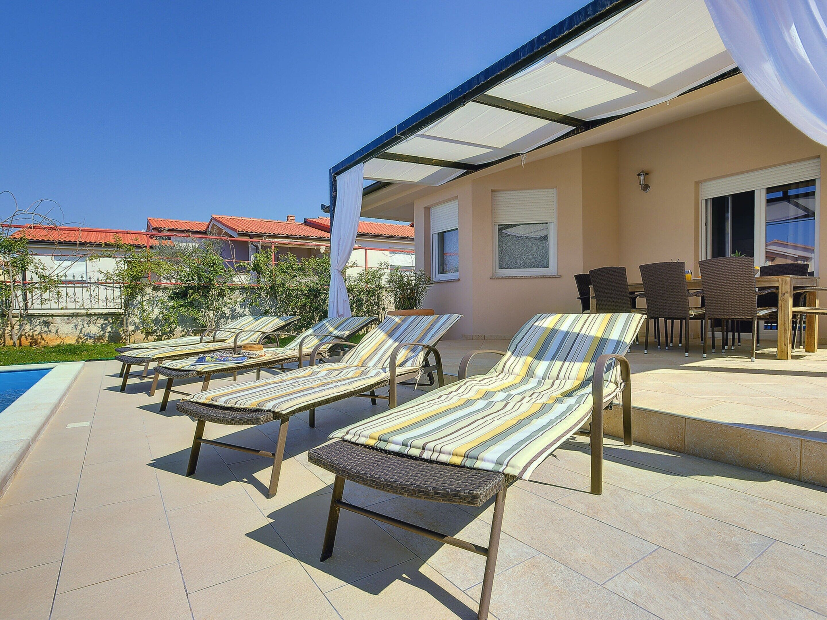 Un espacio de estar al aire libre cubierto con sillones reclinables y una mesa, con vistas a una piscina y edificios cercanos.