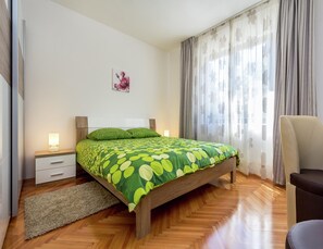A bedroom with a wooden bed, a nightstand with a lamp, a chair, and a window with curtains. - Apartment in Pula Near the Sea and Town Center (Pula)