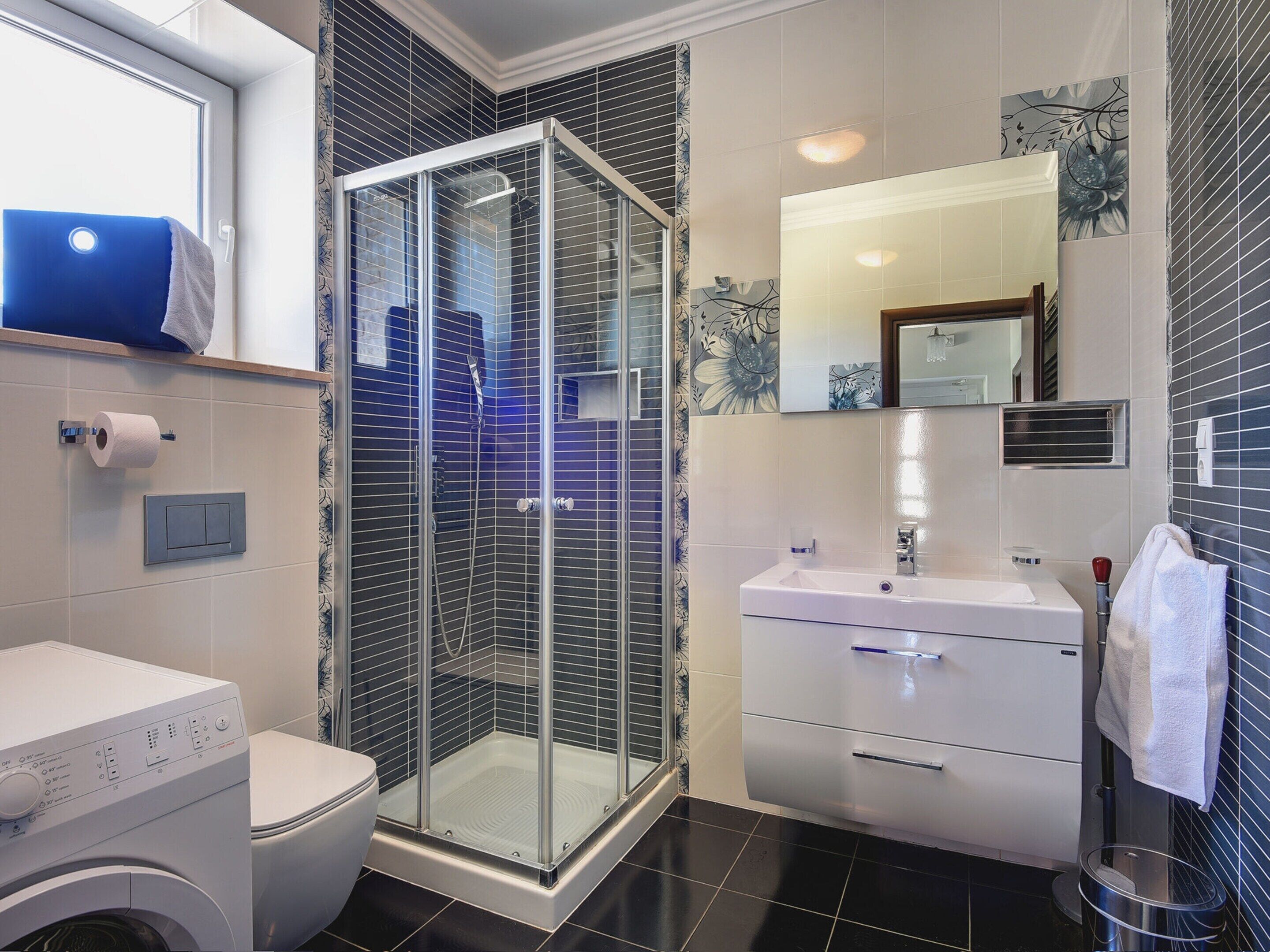 A modern bathroom with a glass-enclosed shower, a toilet, a washing machine, and a sink with a mirror.