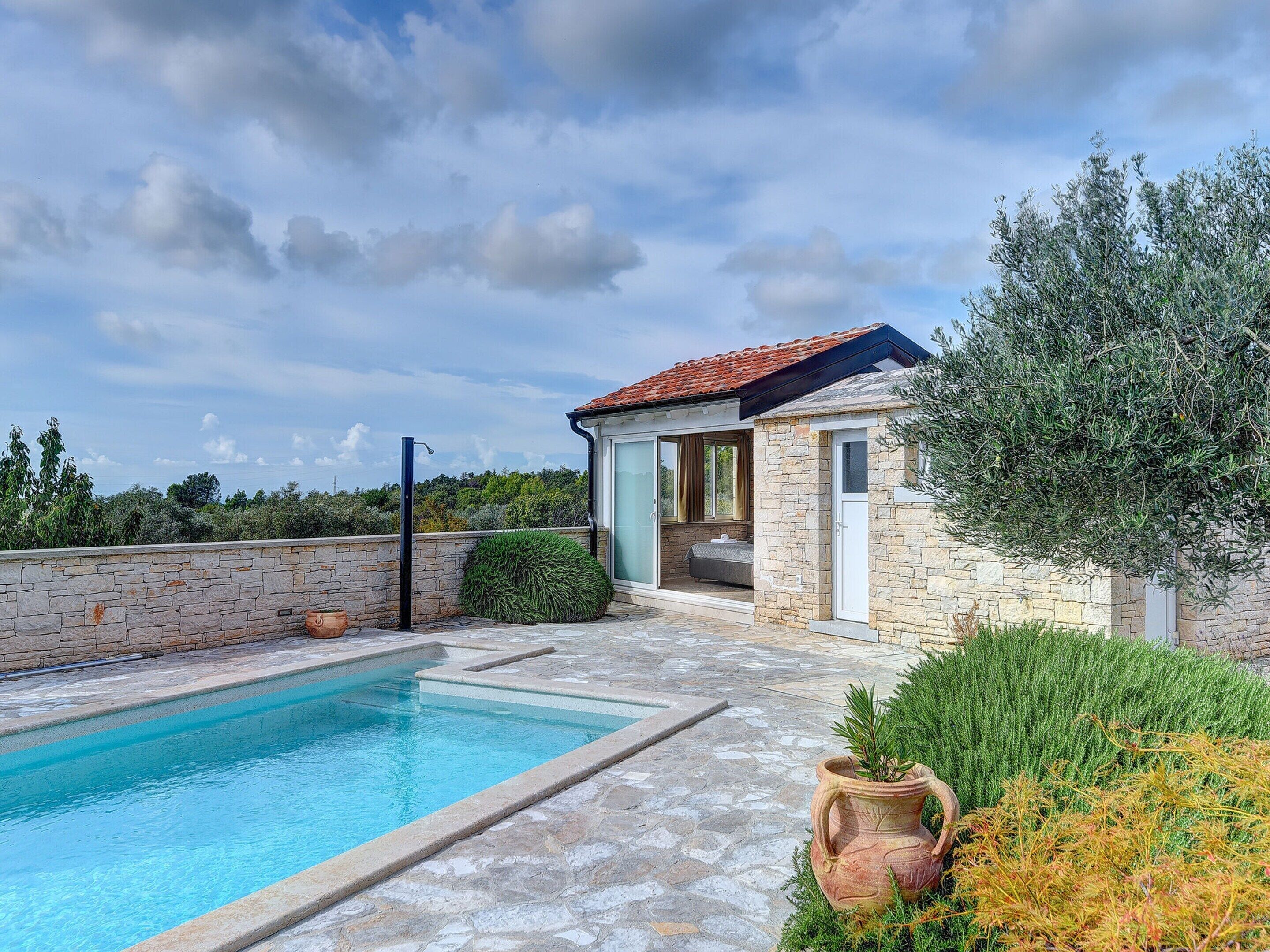 A poolside area with a stone pool, a stone building with a tiled roof, and surrounding greenery.