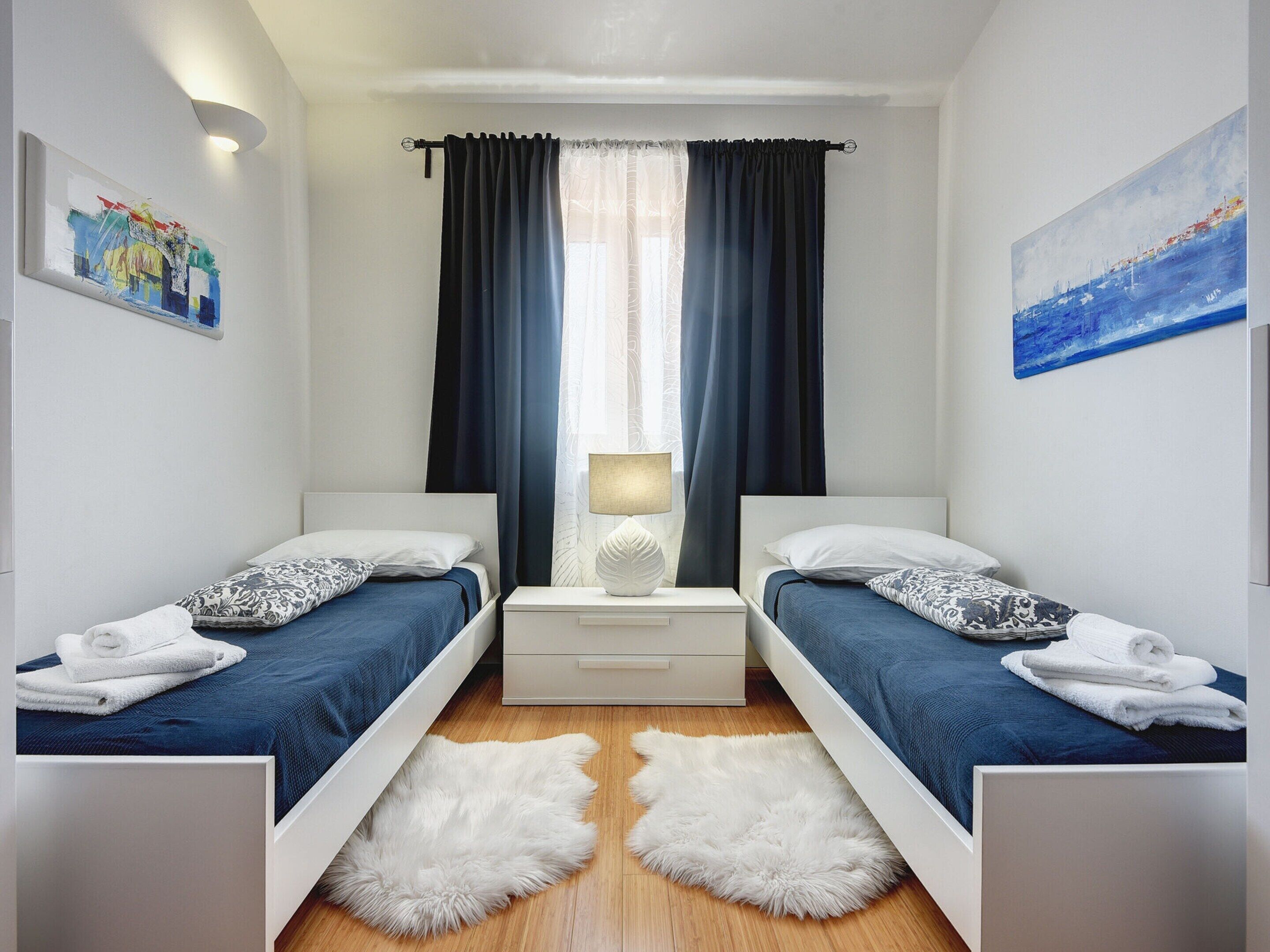 A twin bedroom with two beds, a nightstand with a lamp, a fluffy rug, and artwork on the walls.