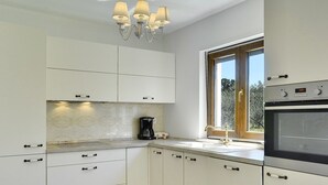 Une cuisine moderne avec des meubles blancs, un four encastré, une machine à café et une fenêtre donnant sur un paysage verdoyant.