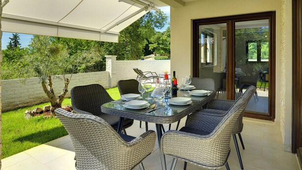 Un espace extérieur aménagé pour les repas, avec des chaises en osier et une table en verre, un barbecue et une vue sur le jardin et les arbres.