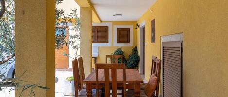 Un patio exterior con una mesa y sillas de madera, rodeado de paredes amarillas y una puerta con contraventanas cerradas.