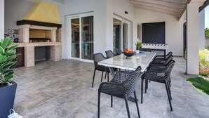 Een terras buiten met een eettafel, stoelen, een barbecue en een glazen schuindeur die naar een binnenruimte leidt.