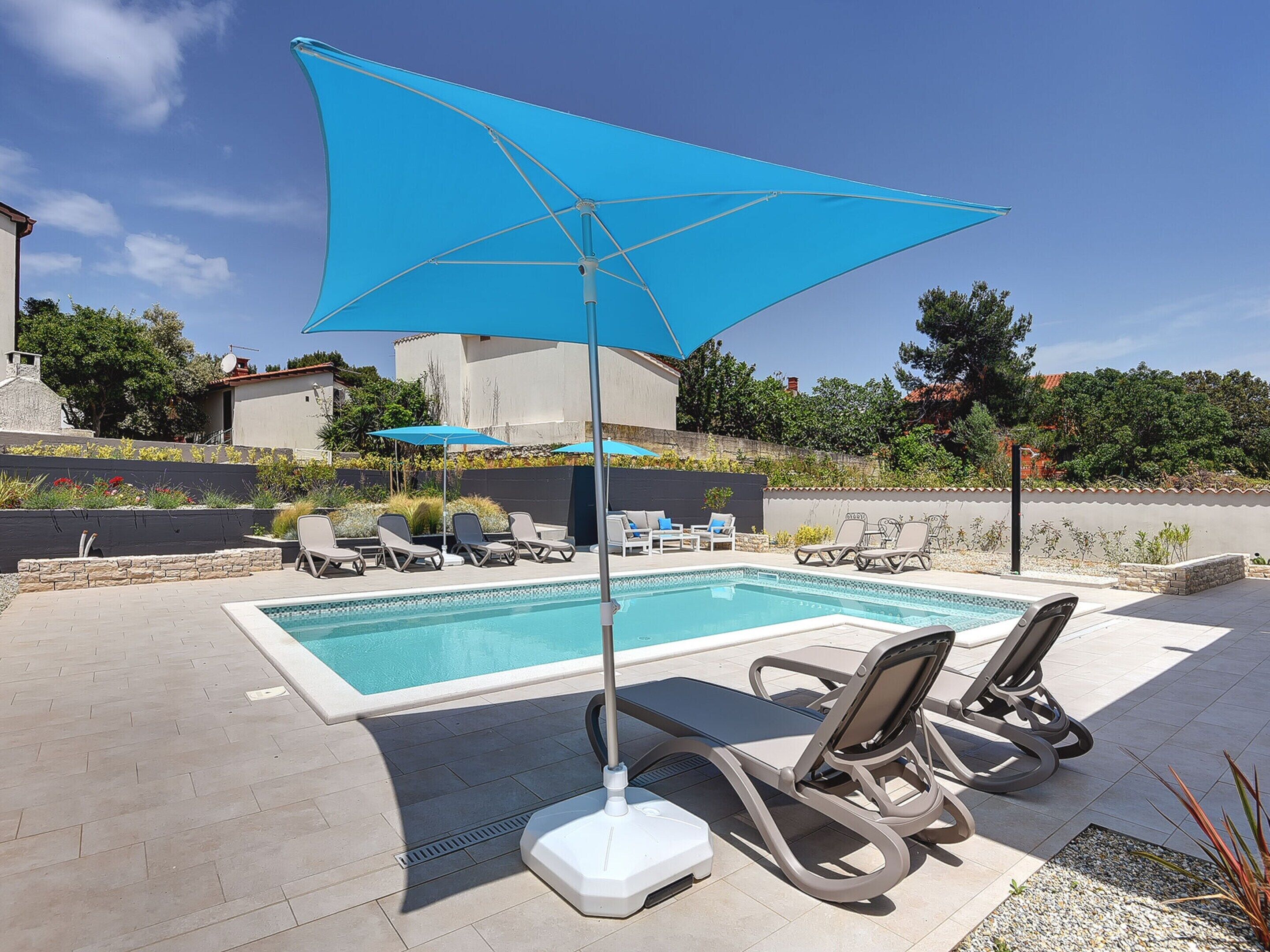 Un área de piscina con una sombrilla azul, sillas de descanso y un cielo despejado.