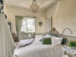 A bedroom with a large bed, a chandelier, a window with curtains, a bedside table, and a mirror. - Magnificent Villa in Groznjan With High-end Kitchen & Pool (Groznjan)