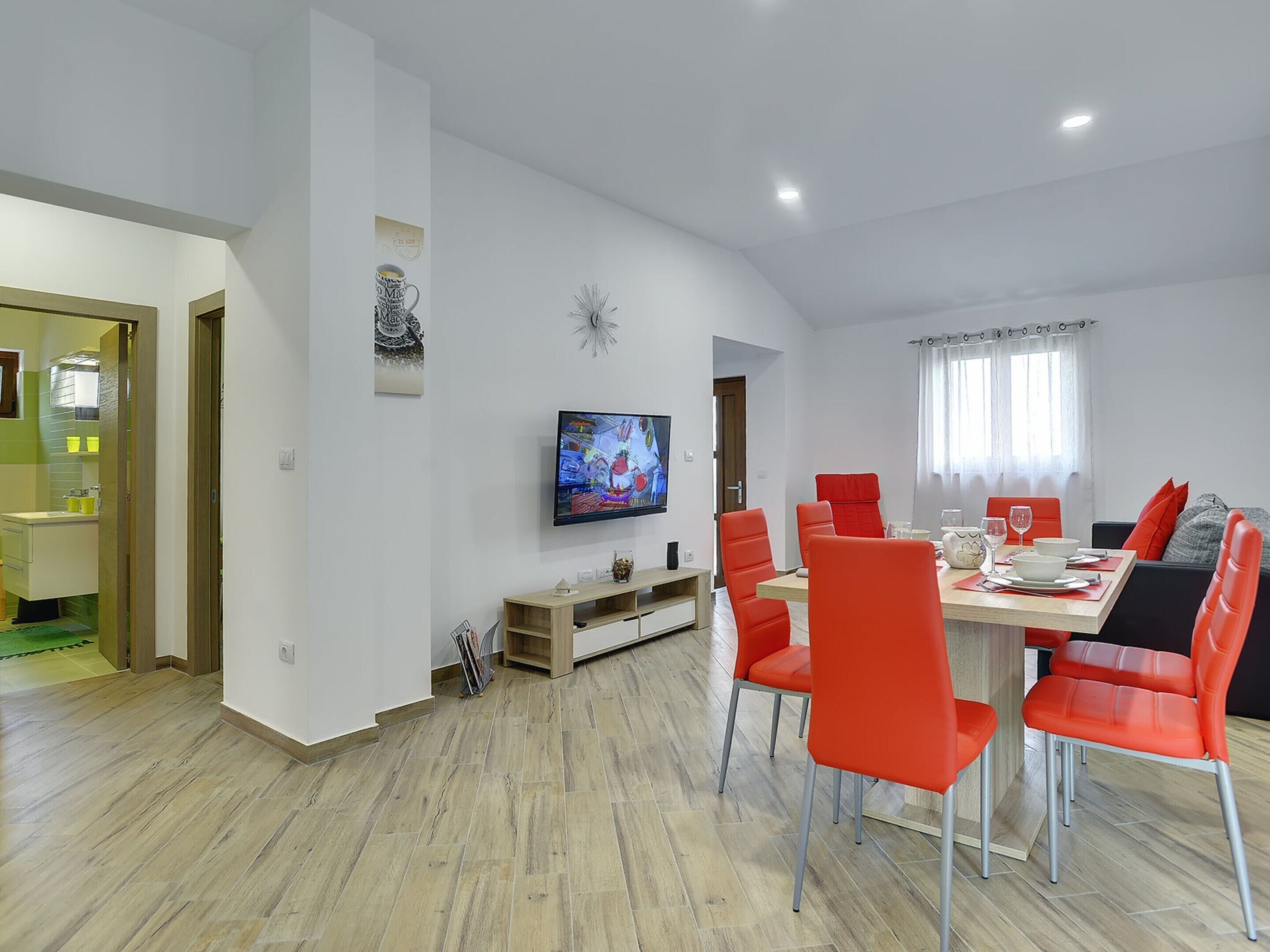 A modern dining area with a wooden table, red chairs, and a flat-screen TV mounted on the wall.