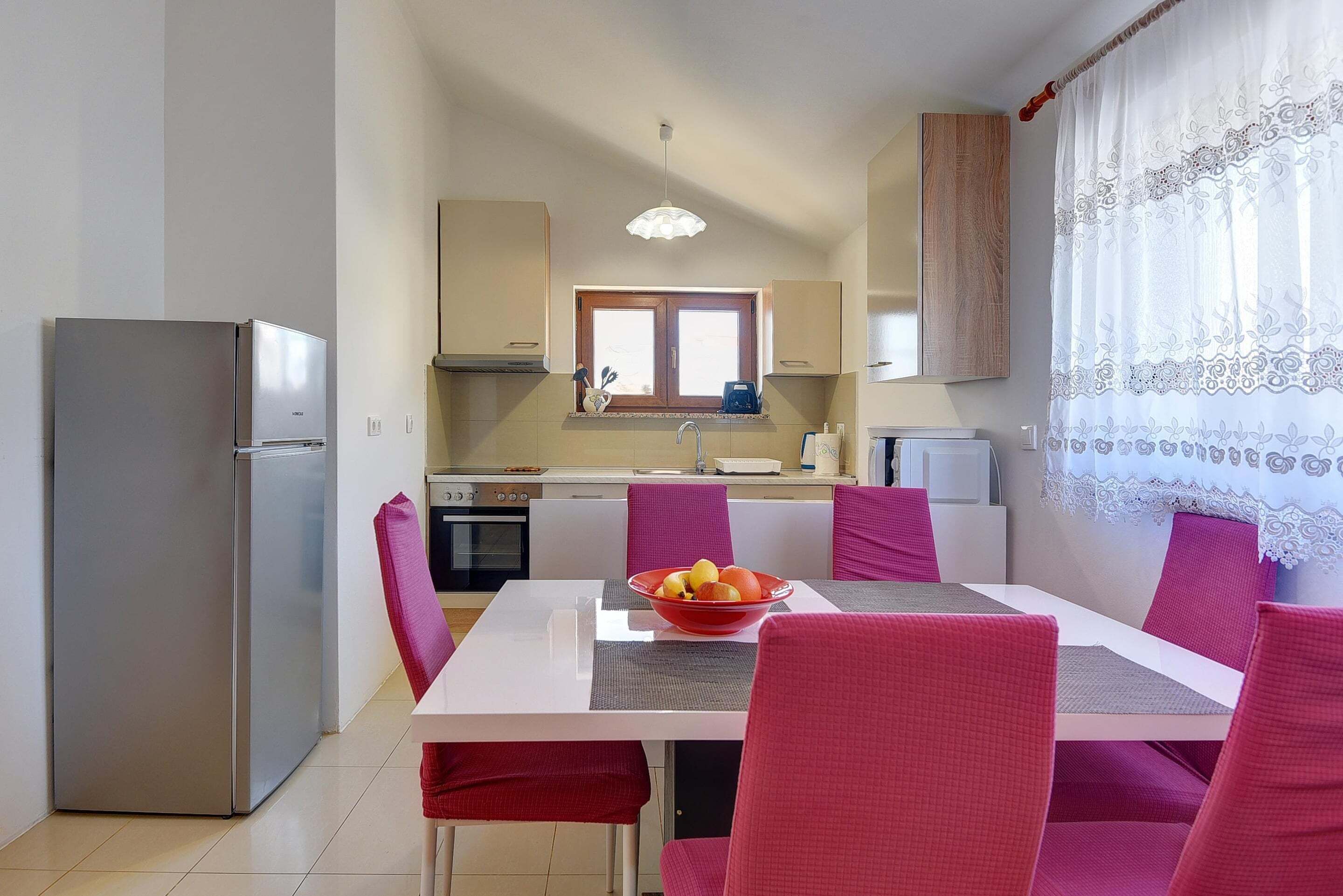 Una cocina compacta con refrigerador, una mesa de comedor con sillas y una ventana con cortinas.