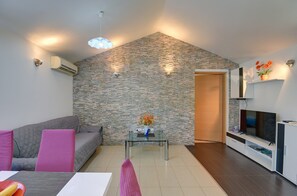 A modern living room with a stone wall, a glass coffee table, a grey sofa, and a TV on a white cabinet. - Holiday Home in Medulin Near Beach (Medulin)