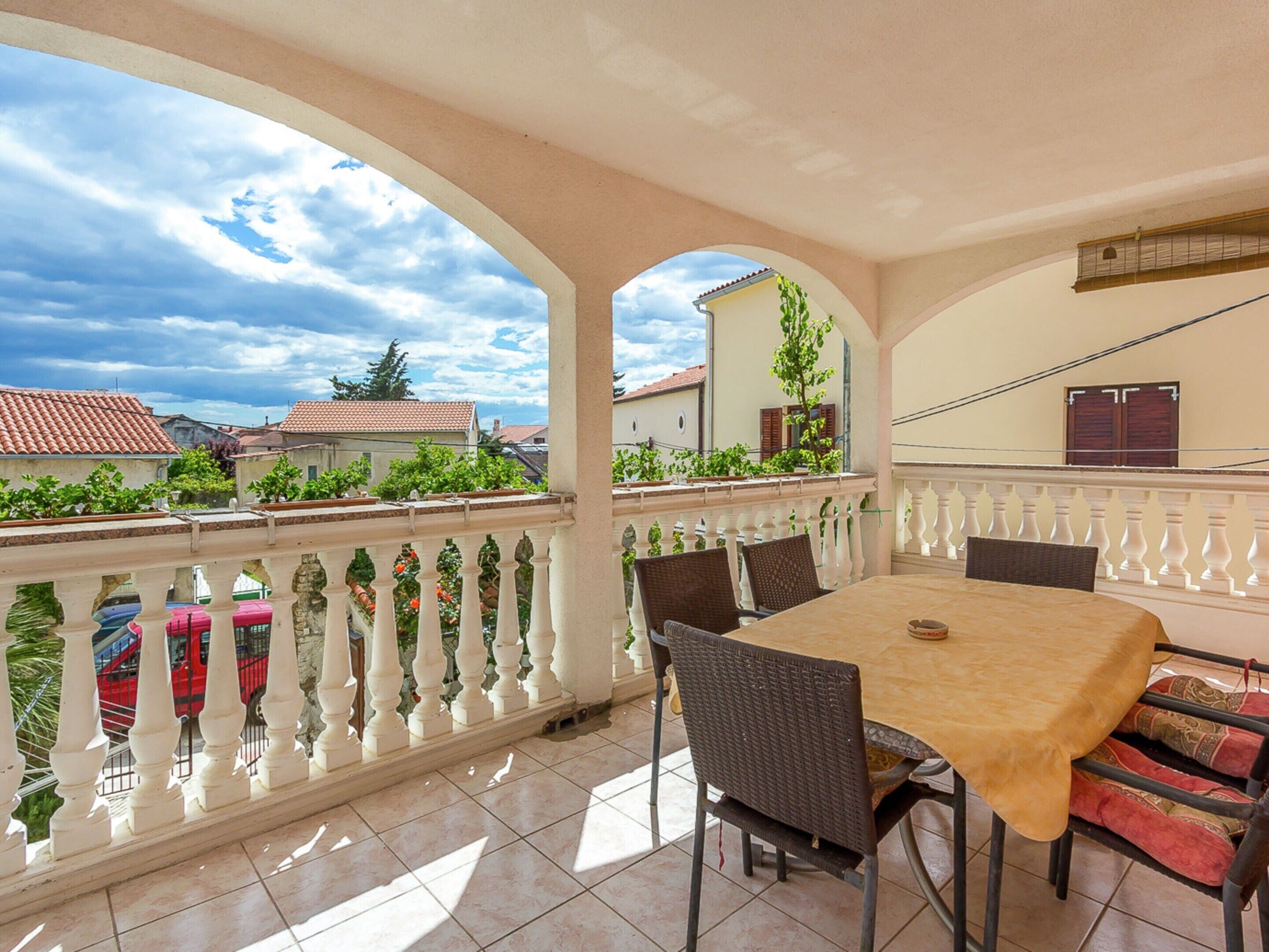 Un patio techado al aire libre con una mesa y sillas, barandilla blanca y vistas al vecindario.