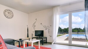 A modern living room with a grey sofa, a flat-screen TV, a small coffee table, and a red rug. A large window with curtains offers a view of a swimming pool and greenery. - Detached Villa With Pool and Large Garden in Quiet Area Near the Sea (Barban)