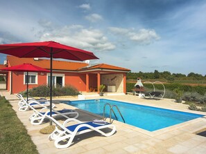A swimming pool with lounge chairs and a red umbrella, a barbecue area, and a view of vineyards.