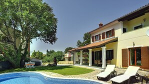 En swimmingpool med liggestole, en overdækket terrasse og en bygning med tegltag.