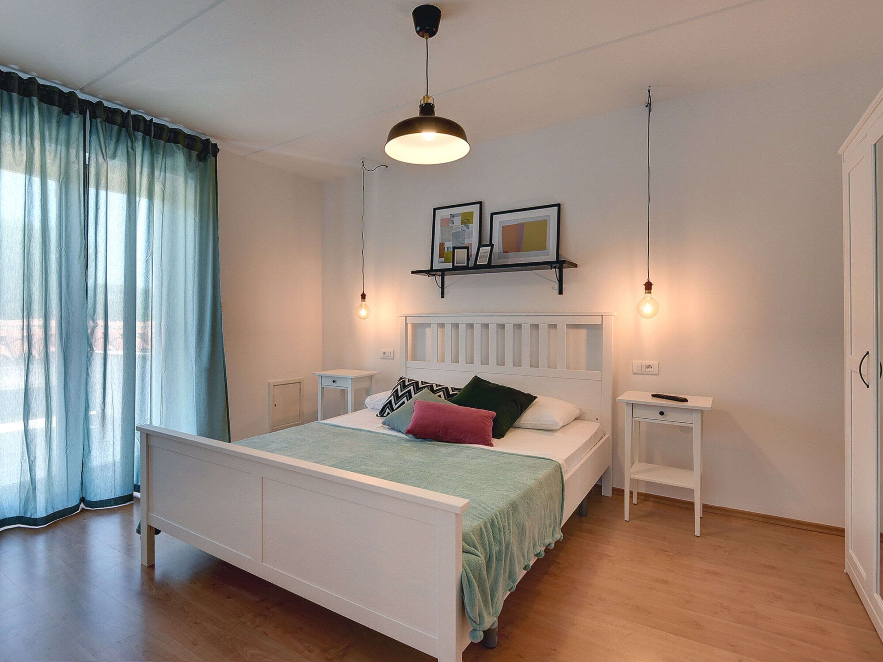 Une chambre à coucher avec un lit blanc, une table de chevet, une commode, un luminaire suspendu et une fenêtre avec des rideaux.